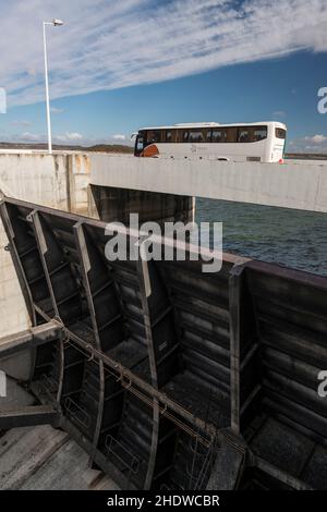 Un autobus turistico che conduce lungo una delle porte d'alluvione sulla cima della diga di Alqueva, la diga più grande e il lago artificiale dell'Europa occidentale vicino Moura, Portogallo Foto Stock