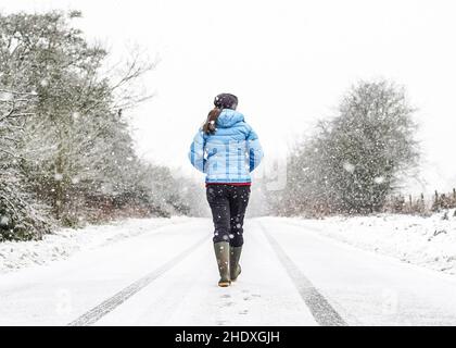 Vista posteriore di una giovane donna con cappotto e pozzi invernali, isolata all'aperto nella campagna del Regno Unito, camminando in mezzo alla corsia di campagna deserta in caduta di neve. Foto Stock