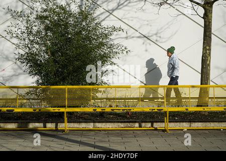 Un uomo con una sigaretta in bocca, con la cenere non ancora caduta, cammina oltre un muro sul quale cade la sua ombra con la sigaretta. Davanti a lui una panca. Foto Stock