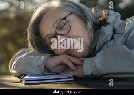 donna più vecchia con capelli bianchi e occhiali addormentati sul suo notebook nel parco sognando Foto Stock