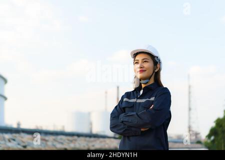Donna asiatica ingegnere braccio attraversato e sorriso con fiducioso in attesa di futuro con la fabbrica di raffineria di petrolio in background. Foto Stock