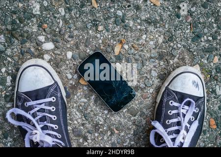 In piedi su uno smartphone rotto in strada. Telefono cellulare danneggiato a terra. Foto Stock