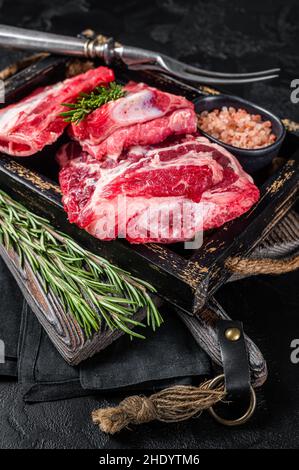 Costolette di manzo organico crudo pronte per essere cuote in vassoio di legno con erbe. Sfondo nero. Vista dall'alto Foto Stock