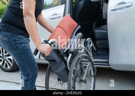 Donna asiatica pieghevole e sollevare la sedia a rotelle nella sua auto. Concetto di accessibilità. Foto Stock