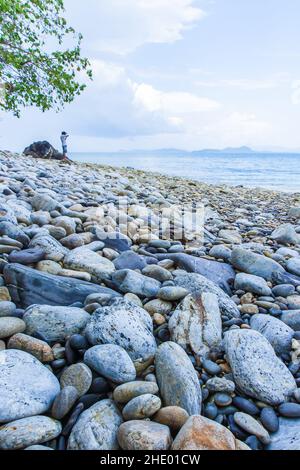 Vista sul mare della spiaggia di ghiaia tropicale in estate soleggiata, un uomo turistico si erge sullo sfondo. Isola di Kang Kao, Ranong, Tailandia. Foto Stock