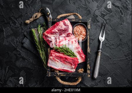 Costolette di manzo organico crudo pronte per essere cuote in vassoio di legno con erbe. Sfondo nero. Vista dall'alto Foto Stock