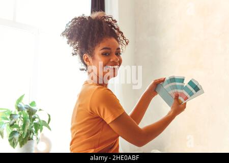 Sorridente ragazza afroamericana si alza di nuovo alla macchina fotografica nel mezzo della camera da letto con tavolozza di sfumature blu per la ristrutturazione degli interni, scegliendo la vernice per bala Foto Stock