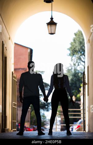 Silhouette umane in arca antico con lampada da strada appesa sopra. L'uomo e la donna si tengono a mano, girati indietro, alberi e case dentro Foto Stock