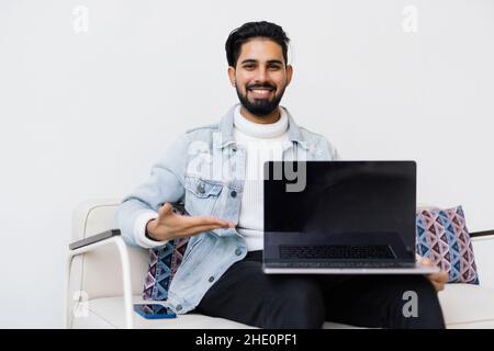 Giovane uomo indiano che si trova comodamente sul divano mentre si utilizza il computer portatile con schermo vuoto. Foto Stock