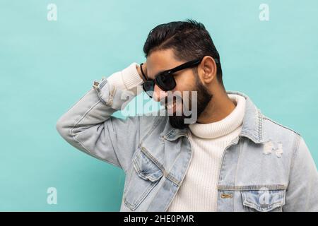 Foto di fiducioso allegro bello uomo indiano estate vacanza indossare occhiali verde cappuccio isolato teal colore sfondo Foto Stock