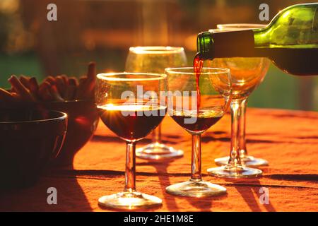 Versare il vino rosso dalla bottiglia in bicchieri sul tavolo con spuntini sullo sfondo durante il tramonto Foto Stock