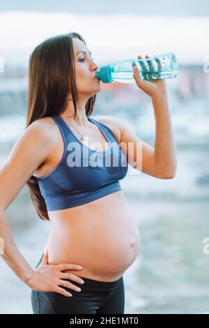 Giovane donna incinta che indossa il reggiseno sportivo bere acqua dopo l'allenamento. Reidratazione, idroequilibrio, reintegro, deficit idrico. Foto Stock