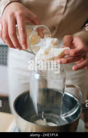 Colpo tagliato verticale di artigiano femminile che aggiunge cera di soia secca bianca in vaso di vetro in acqua bollente per creare la miscela di costruzione di candela. Foto Stock