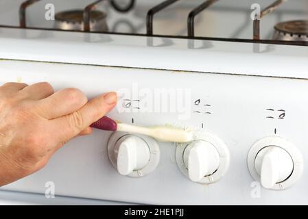 La donna lava a mano i rubinetti della stufa a gas bianca con uno spazzolino. Un modo insolito di pulizia della casa Foto Stock