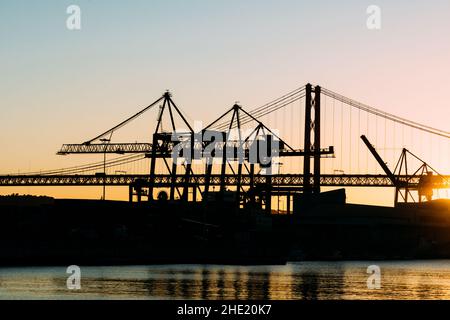 Silhouette di gru al porto di Lisbona, Portogallo, con il ponte del 25 aprile sullo sfondo al tramonto Foto Stock