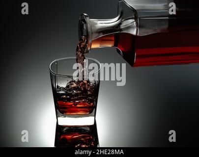 Il whisky viene versato da un decanter in un bicchiere congelato con ghiaccio naturale. Sfondo nero riflettente. Foto Stock