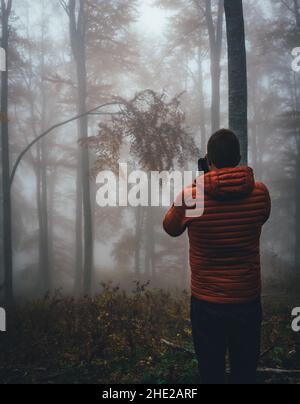Giovane escursionista portatore con giacca arancione che scatta foto alla foresta con nebbia e nebbia. Vista posteriore del fotografo sul campo godendo la vista. Foto Stock