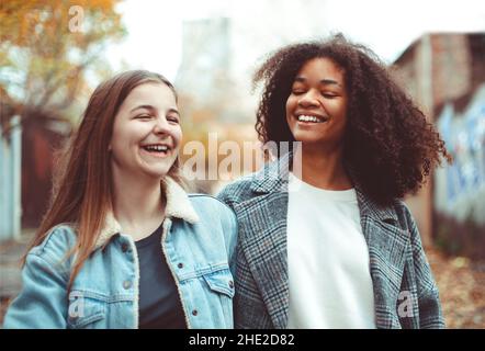 Due allegre fidanzate multirazziali che camminano per la strada della città, parlano e condividono storie di vita, le migliori amiche multietniche che si godono a piedi autunno Foto Stock