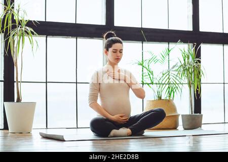 Meditazione durante la gravidanza. Giovane calma tranquilla donna incinta che fa yoga a casa, meditando e praticando la consapevolezza, madre in attesa seduta Foto Stock