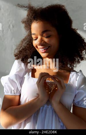 Giovane pacifica riconoscente donna afroamericana con occhi chiusi in abito bianco che fa segno di cuore, luci a LED in mani di female nero tranquillo Foto Stock