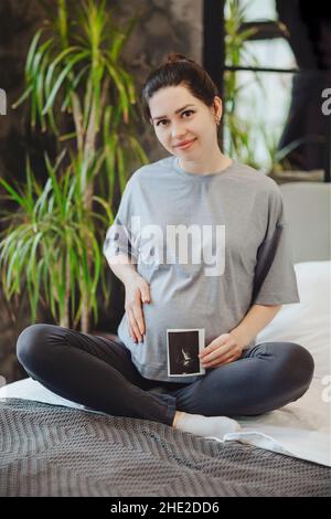 Donna incinta che tiene la gravidanza ultrasuoni mentre si siede in posa di lotus sul letto a casa, gravidanza madre con sonogramma bambino. Gravidanza, maternità e. Foto Stock