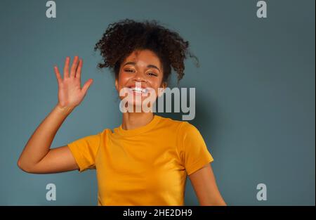 Bella ragazza di etnia afroamericana felice scuro con pelle in maglia gialla che alza la mano nel saluto mentre guarda in macchina fotografica con piacevole sorriso, p Foto Stock