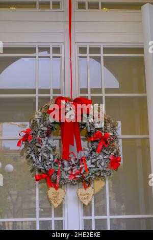 Corona di Natale con ciotole rosse appese al primo piano della porta bianca. Decorazione esterna di Natale Foto Stock