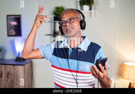 Uomo anziano attivo con cuffie che ballano ascoltando musica sul telefono cellulare a casa - concetto di stile di vita sano, gioiosa e rilassante Foto Stock