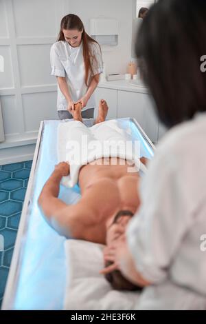 I terapisti del massaggio lavorano con le gambe e la testa di un paziente di sesso maschile che giacciono su un comodo divano incandescente Foto Stock