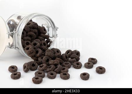 anelli di mais per la colazione, gustosi anelli neri e cereali versati da un vaso di vetro su sfondo bianco, concetto cibo sano Foto Stock