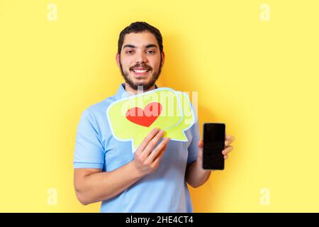 Il giovane uomo tiene in mano un fumetto con un cuore e mostra lo schermo vuoto dello smartphone. Concetto di social media. Foto Stock