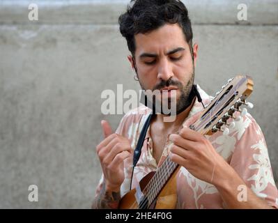 Bel giovane latino-americano con barba nera e maschera facciale sotto il mento suona una piccola chitarra andina (charango) con dieci corde. Foto Stock