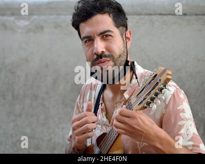 Bel giovane latino-americano con barba nera e maschera facciale sotto il mento suona una piccola chitarra andina (charango) con dieci corde. Foto Stock