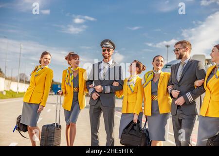 Donne gioiose stewardesses che tengono borse di viaggio e sorridenti mentre si trova in aeroporto con piloti maschi Foto Stock