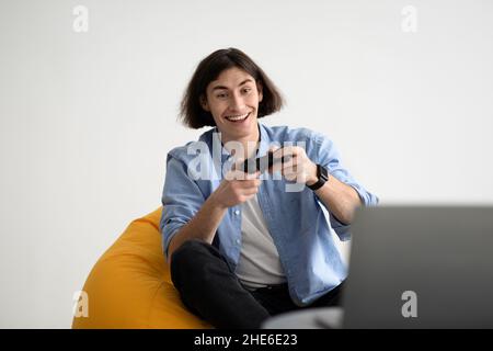 Ragazzo emozionale giovane che gioca video gioco con joystick a casa, seduta poltrona beanbag, spazio copia Foto Stock