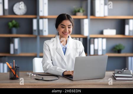 Sorridente giovane cute indiana donna medico lavoratore, medico in uniforme digitando sul computer in ufficio interno Foto Stock