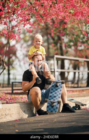 Ragazzino seduto sulle spalle del padre seduto vicino allo skateboard nel parco. Buon padre e figlio divertirsi dopo lo skateboard. Padri da Foto Stock