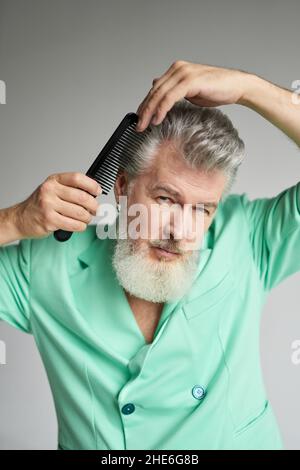 Ritratto di bell'uomo di mezza età con barba che guarda la macchina fotografica, spazzolando i capelli con il pettine, posando su sfondo bianco Foto Stock