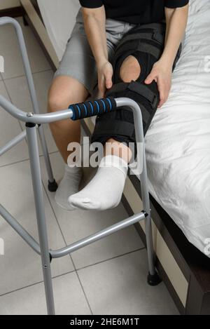 Donna che indossa un tutore per ginocchio regolabile dopo la frattura della gamba seduta sul letto cercando di alzarsi con il camminatore. Recupero e riabilitazione dopo intervento chirurgico Foto Stock