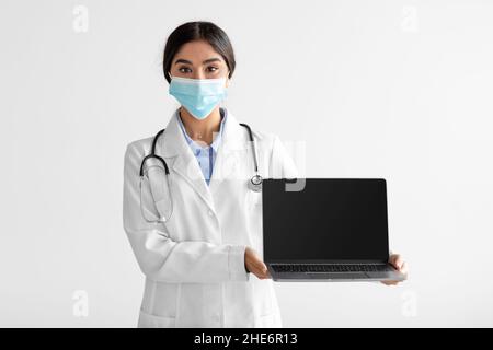 Giovane attraente medico indù in uniforme e maschera protettiva mostra computer con schermo vuoto Foto Stock