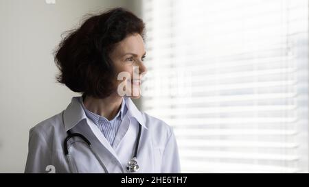 Sognante sorridente bella ragazza medico GP guardando fuori dalla finestra. Foto Stock