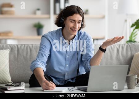 Ragazzo che indossa le cuffie e che ha una lezione online sul portatile, iscritto informazioni, seduto sul divano a casa Foto Stock