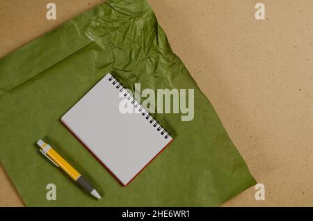 Un notebook aperto con fogli bianchi vuoti e una penna gialla contro una superficie multicolore. Carta verde sgualcita su sfondo beige. Vista dall'alto. S Foto Stock