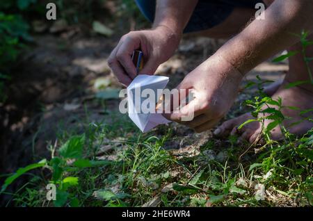 Un uomo adulto a piedi nudi illumina una barca di carta. Mani che tengono una barca bianca per bambini origami e un fiammifero ardente. Ora legale. Trick selettivo. All'esterno del Foto Stock