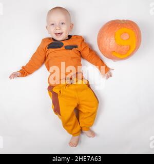 Sorridente neonato felice nove mesi bambino nei vestiti arancioni sul letto luminoso e arioso copia spazio infanzia con zucca con il numero 9 scolpito su di esso Foto Stock