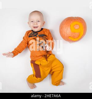 Novosibirsk, Russia - 11. 10. 2021: Sorridente neonato felice nove mesi in abiti arancioni a letto, infanzia con zucca con il numero Foto Stock