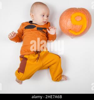 Sorridente neonato felice nove mesi bambino nei vestiti arancioni sul letto luminoso e arioso copia spazio infanzia con zucca con il numero 9 scolpito su di esso Foto Stock