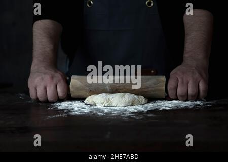 Man Baker o chef sta arrotolando l'impasto fresco con un rullo su una superficie di legno. Foto Stock