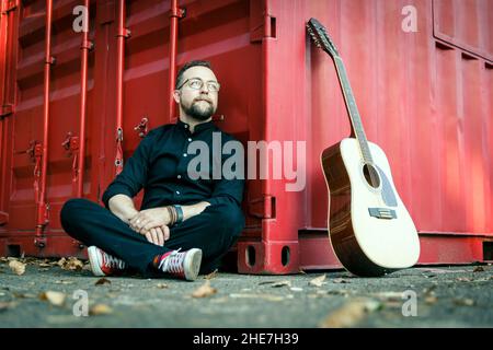 Uomo in nero con una barba seduta su un terreno sotto un contenitore rosso con chitarra acustica a 12 corde Foto Stock