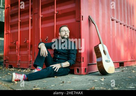 Uomo in nero con una barba seduta su un terreno sotto un contenitore rosso con chitarra acustica a 12 corde Foto Stock